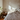 A cozy bedroom with a unmade bed, featuring white bedding and pillows. Sunlight streams through a window, casting patterns on the textured wall. A potted plant and stack of books on a wooden nightstand add to the relaxed, lived-in atmosphere.