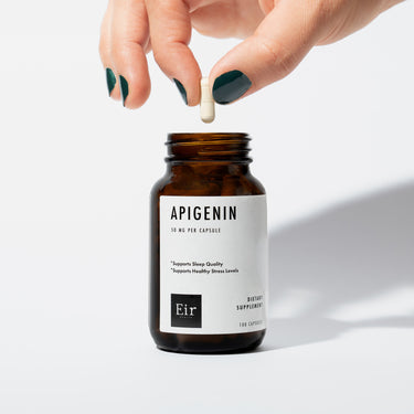 Close-up of a hand with dark green nail polish holding a white Apigenin capsule above an open bottle of Eir Apigenin supplement. The bottle's label is visible, showing product information and benefits.