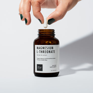 Close-up of a hand with dark green nail polish holding a white capsule above an open bottle of Eir Magnesium L-Threonate supplement. The bottle's label is visible, showing product information.