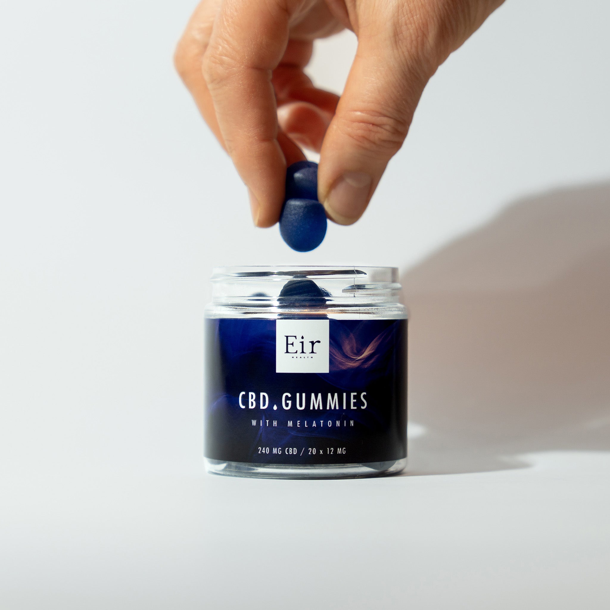 Dłoń wyjmująca niebieskiego żelka CBD z przezroczystego słoika z etykietą Eir CBD Gummies z melatoniną, 240 mg CBD, na białym tle.