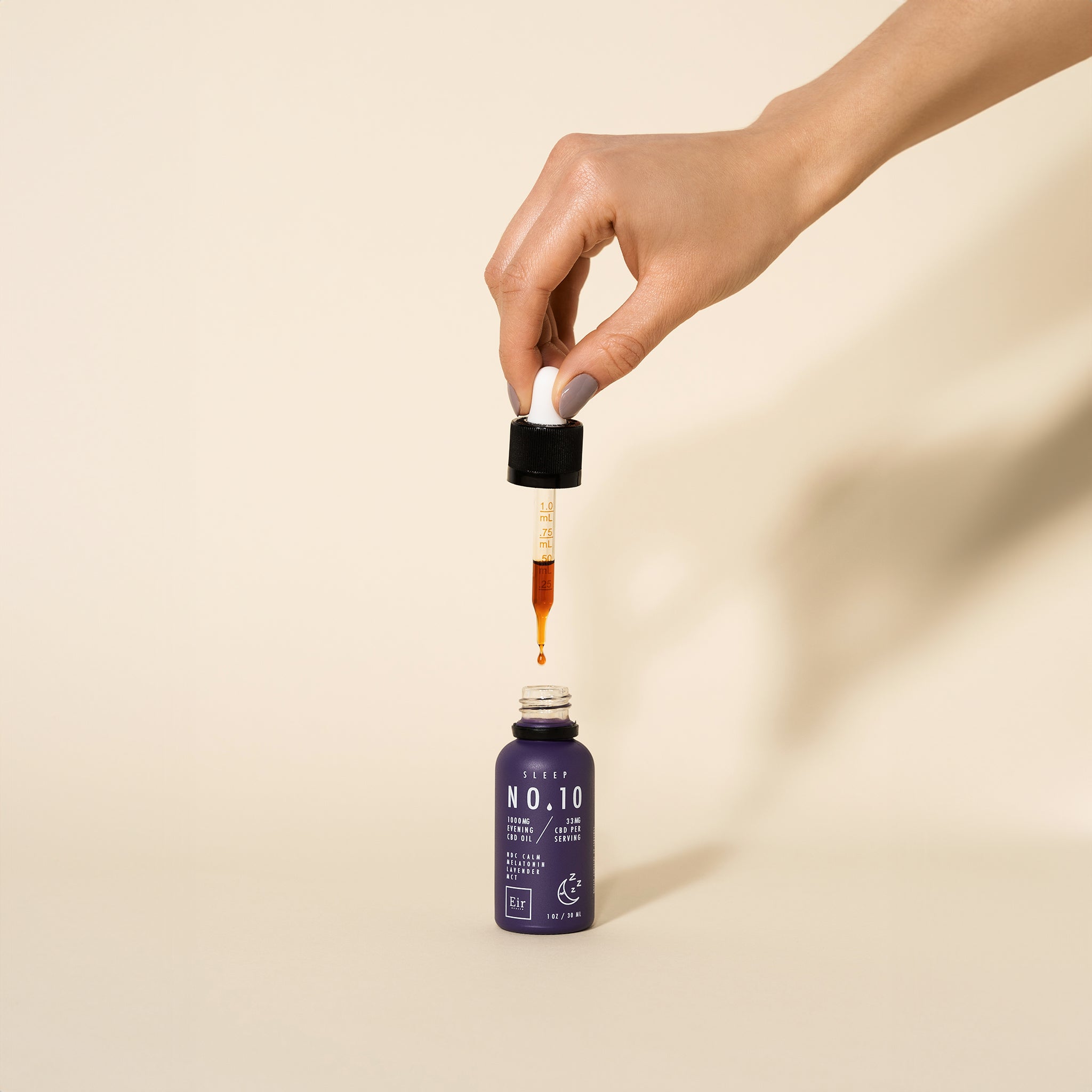 Hand holding a dropper above a purple bottle of Eir No. 10 CBD Oil, with a drop of oil suspended at the end, against a neutral background.