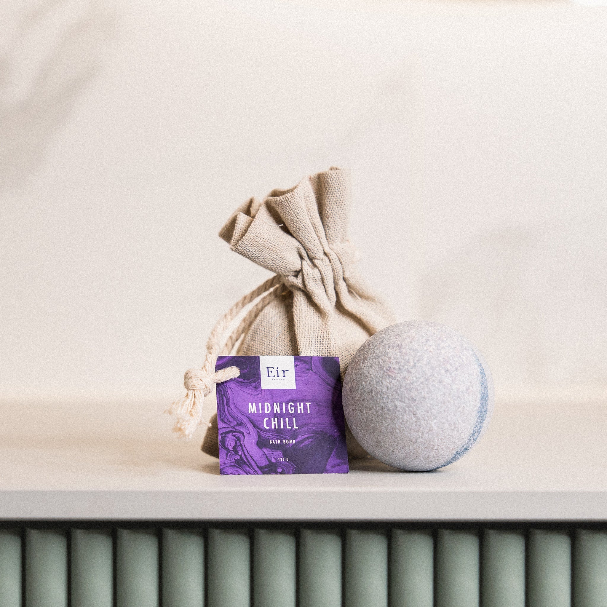  Packaging of Eir's 'Midnight Chill' bath bomb, a jute pouch, and a grey bath bomb on a radiator in a bright interior.