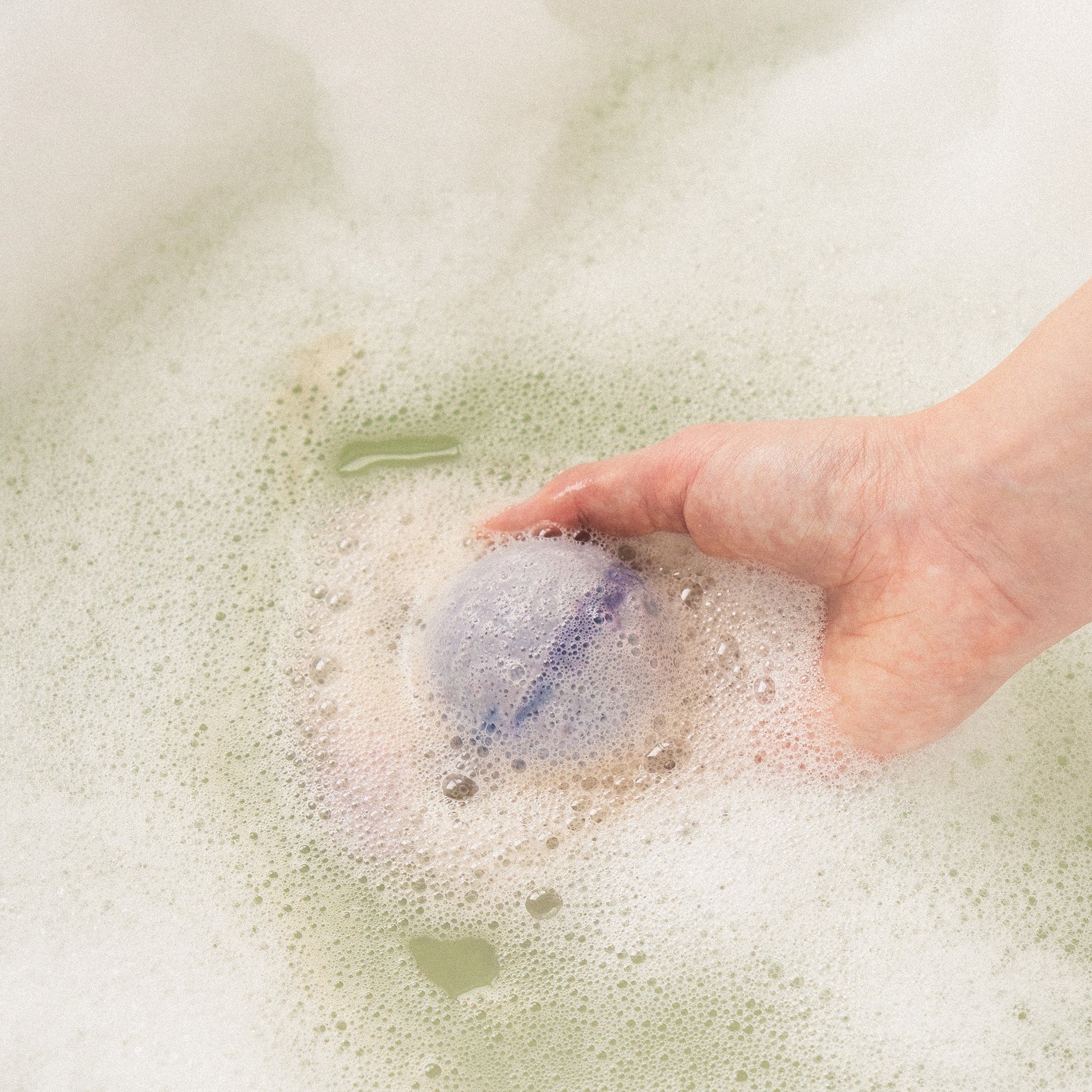 Hand holding Eir's 'Midnight Chill' grey bath bomb in bathtub foam, creating a relaxing atmosphere.