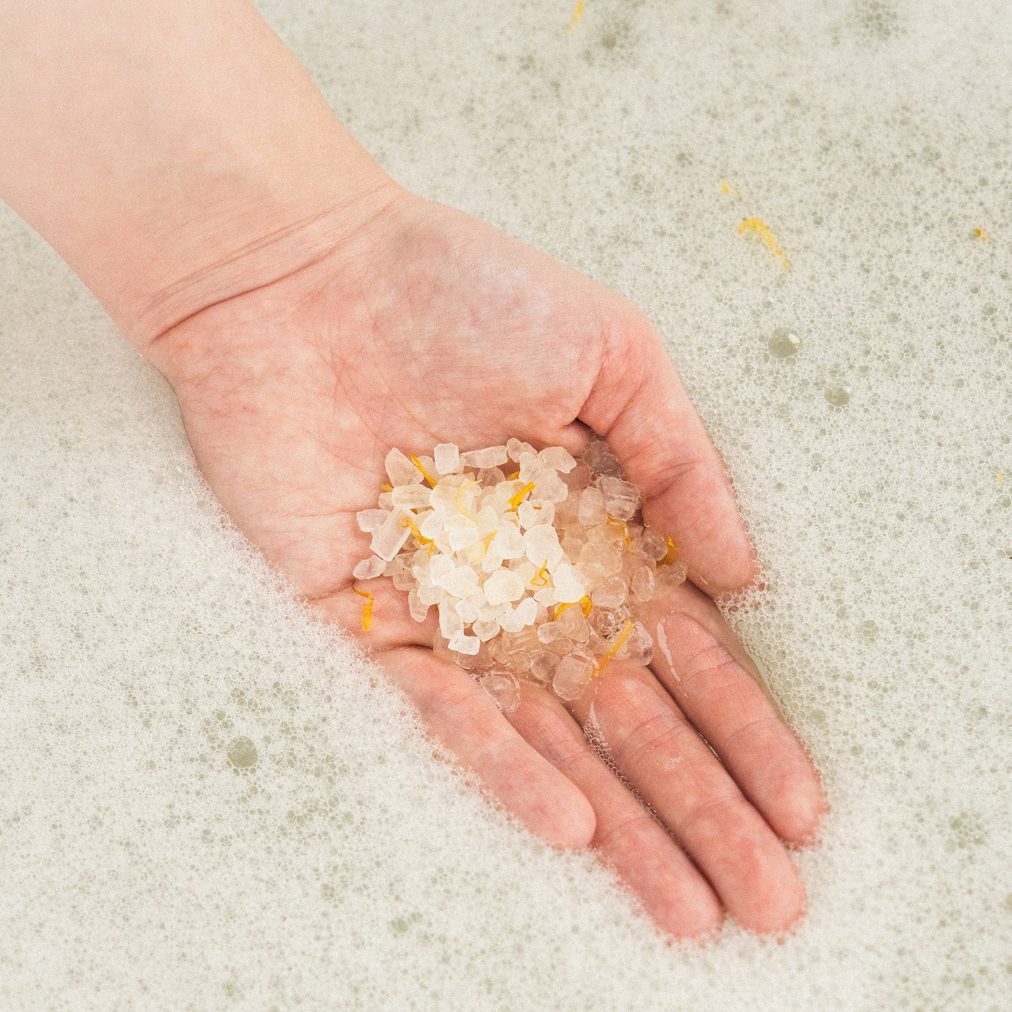 A hand holding transparent bath salt crystals with golden flakes, against a background of white, foamy water.