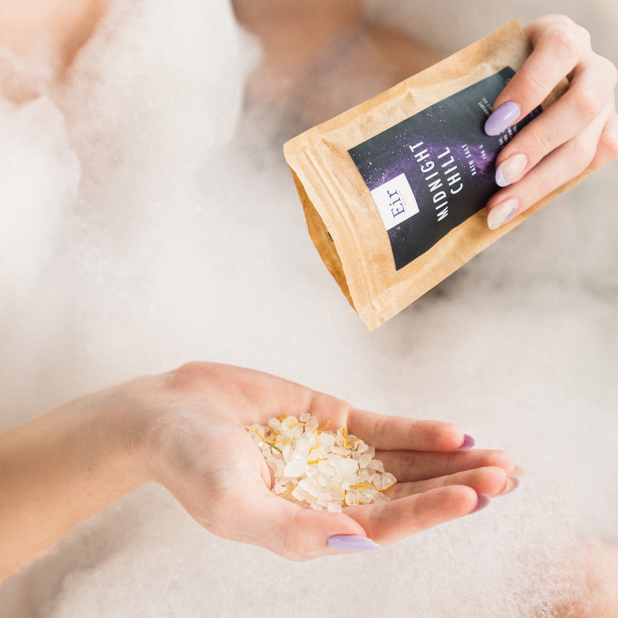 A woman's hand with light-colored nails pouring white bath salt crystals from the paper packaging of "Midnight Chill."